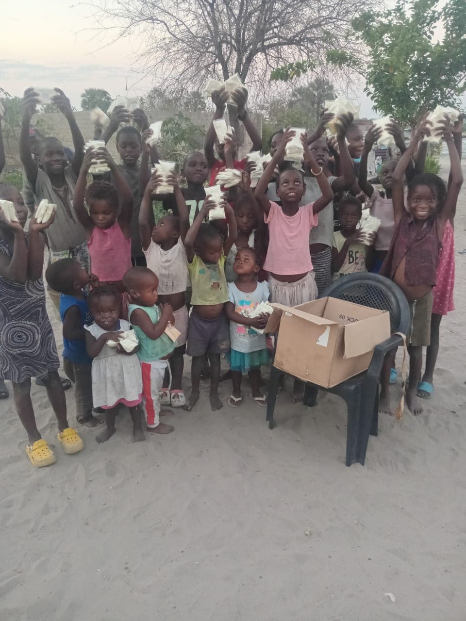 Children gathered outdoors holding packets of food beside a box of supplies.