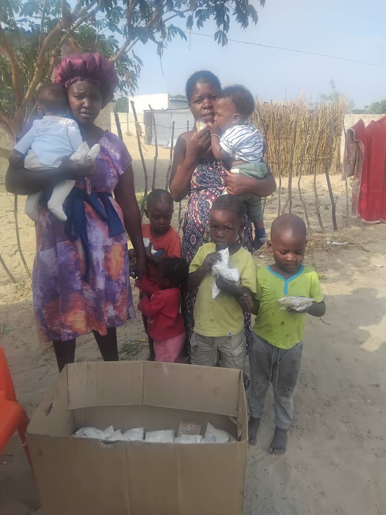 Caregivers and children standing together beside a box of food packets.