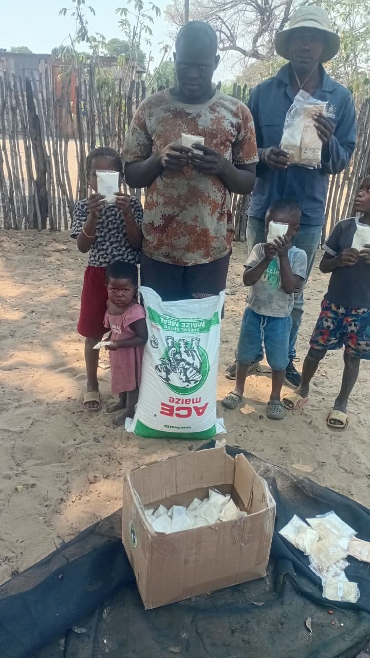 Community members and children with food packets, a bag of maize meal, and a box of supplies.